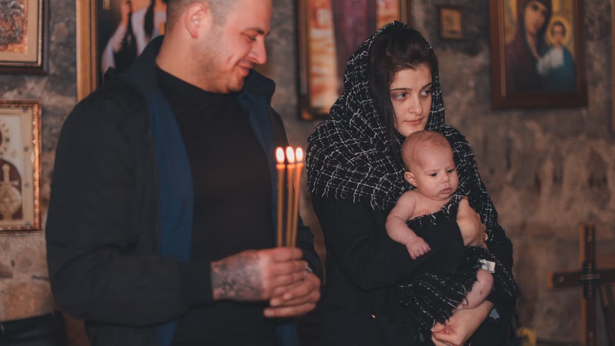 A baby held by mother during an Orthodox candle ceremony indoors with religious icons.
